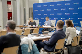 Pokaż zdjęcie: Kobieta i mężczyzna siedzą za stołem. Kobieta mówi do mikrofonu.