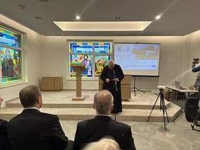 Pokaż zdjęcie: Ksiądz w czarnej sutannie stoi pochylony przed  mikrofonem. Przed nim wśród widowni dwaj mężczyźni ubrani w garnitury i białe koszule. Za księdzem duży witraż z mężczyzną i napisem święty Łukasz.