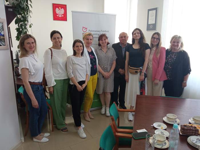 Pokaż zdjęcie: W pokoju na tle baneru z logo PFRON pozuje do pamiątkowego zdjęcia 9 osób, są to dyrekcja oddziału lubelskiego PFRON oraz goście, młode kobiety z Ukrainy