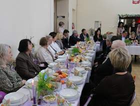 Przy świątecznym stole siedzi bardzo dużo osób. Na stole leżą wielkanocne potrawy i dekoracje. Wszyscy są odświętnie i elegancko ubrani. 