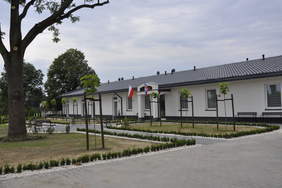 Pokaż zdjęcie: Parterowy, rozłożysty budynek. Nad głównym wejściem wiszą dwie flagi. Przed budynkiem skwer z alejkami i drzewami.