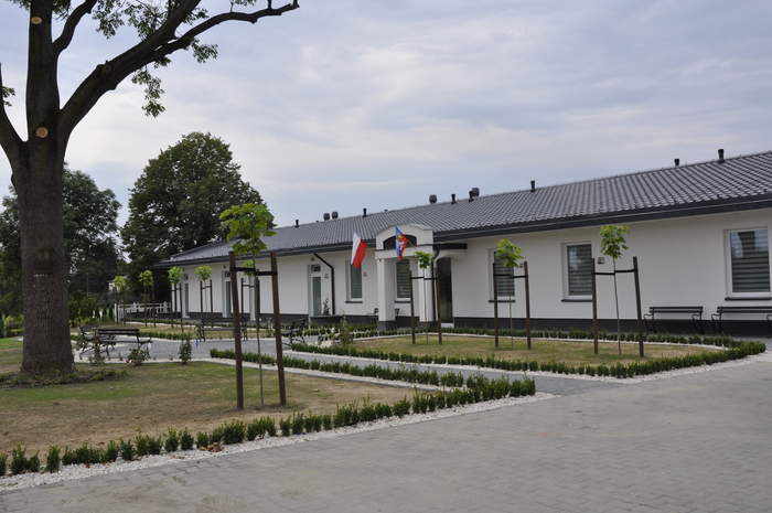 Pokaż zdjęcie: Parterowy, rozłożysty budynek. Nad głównym wejściem wiszą dwie flagi. Przed budynkiem skwer z alejkami i drzewami.