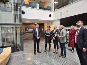 Pokaż zdjęcie: Delegacja w holu Urzędu ogląda nowy budynek. Z lewej strony przeszklony szyb windowy, w tle napis Pomrze Zachodnie 