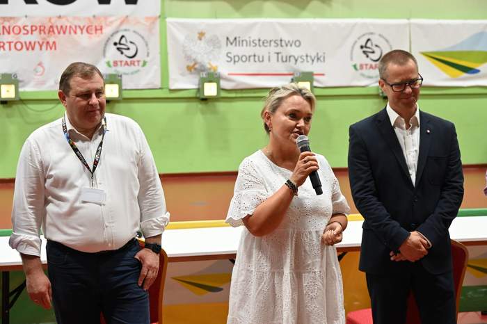Pokaż zdjęcie: Kobieta ubrana w białą sukienkę, z mikrofonem w ręku, w trakcie przemówienia, a za nią dwóch mężczyzn ubranych w koszulę i marynarkę