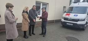 Pokaż zdjęcie: Na zewnątrz przy białym busie udekorowanym kokardą stoją cztery osoby, Dwóch panów przekazuje sobie klucze.