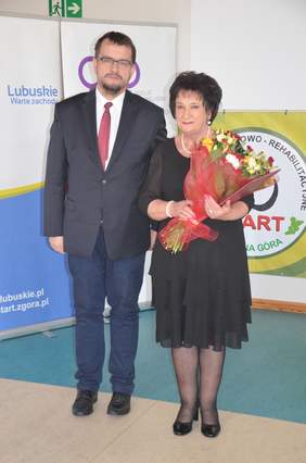 Pokaż zdjęcie: Dyrektor Oddziału Lubuskiego Andrzej Gonia stoi obok Prezes "Start" Zielona Góra Danuty Tarnawskiej na tle loga "Start". Dyrektor jest ubrany odświętnie, a Pani Danuta jest ubrana na czarno i trzyma w rękach kwiaty.