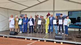 Pokaż zdjęcie: Duża scena plenerowa. Na scenie stoi dużo osób - mężczyzn i kobiet elegancko ubranych. Trzymają dyplomy oprawione w ramy.