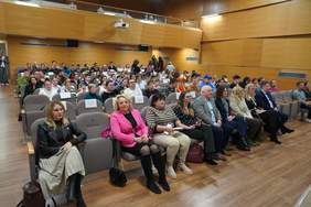 Pokaż zdjęcie: Zdjęcie przedstawia aulę wypełnioną siedzącą publicznością. Aula ma drewniane ściany z rzędami szarych foteli. Wiele osób siedzi zwróconych w stronę sceny. Na pierwszym planie, w przednim rzędzie, siedzą kobiety w jasnych i ciemnych ubraniach. Pod sufitem sali widoczny jest projektor