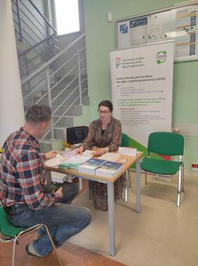 Pokaż zdjęcie: Kobieta reprezentująca PFRON rozmawia przy stole z mężczyzną podczas wydarzenia informacyjnego. Na stole znajdują się ulotki i broszury. W tle widoczny jest banery z logo PFRON i CIDON.