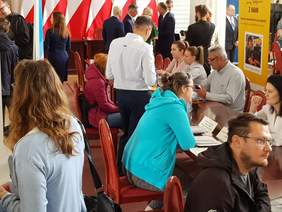 Uczestnicy wydarzenia, część siedzi przy stołach, inni stoją, chodzą