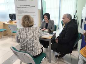 Pokaż zdjęcie: Przy stole siedzą trzy osoby. W tle stoi rollup z logo CIDON.