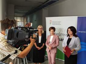 Pokaż zdjęcie: Trzy osoby stoją na tle planszy z logo PFRON-u. Z lewej strony widoczny operator kamery telewizyjnej