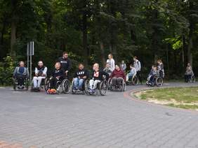 Pokaż zdjęcie: na zdjęciu: miejsce aleje w ośrodku przygotowań olimpijskich , w tle zielone drzewa, 11 osób poruszających się na wózkach jadą na zajęcia objęte programem rehabilitacji. Przy osobach na wózku stoją instruktorzy  