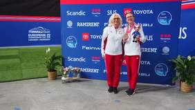 dwie kobiety ubrane w barwach narodowych, zawodniczka trzyma złoty medal uśmiecha się obok niej jej asystentka