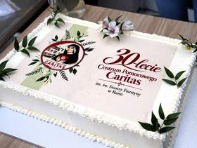 Biały tort jubileuszowy z napisem „30 lat Centrum Pomocowego Caritas”, ozdobiony zielonymi gałązkami, kwiatami i okrągłą grafiką. Tort ustawiony jest na stole.