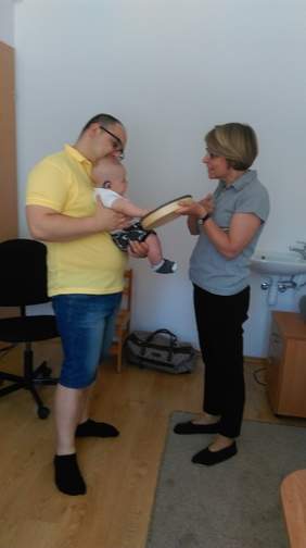 Pokaż zdjęcie: Mężczyzna na stojąco trzyma dziecko, naprzeciwko stoi kobieta z bębenkiem w ręku. 