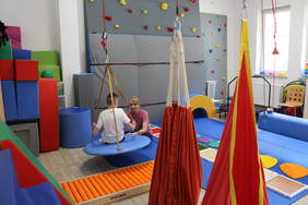 Pokaż zdjęcie: W sali wyłożonej kolorowymi materacami siedzi na hustawce zawieszonej do sufitu chłopiec oboki nieg terapeutka