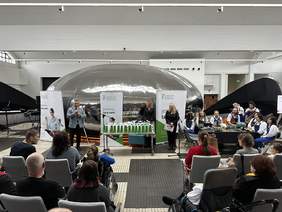 Pokaż zdjęcie: Po lewej stronie Dyrektor Oddziału Pomorskiego podczas przemówienia na tle banerów PFRON oraz srebrej kuli stojacej na środku. Na środku duży stół z nagrodami. Uczestnicy siedzą na szarych krzesłach. 