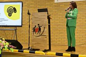 Pokaż zdjęcie: Kobieta w zielonym garniturze, która stoi na scenie i trzyma mikrofon. Prowadzi prezentację, a obok niej na ekranie wyświetlony jest slajd z grafiką ucha oraz napisem dotyczącym badań słuchu. Na mównicy widoczny jest symbol przedstawiający postacie trzymające serce. Całość odbywa się na tle żółtej, ceglanej ściany.