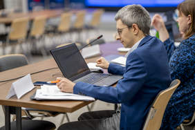 Pokaż zdjęcie: Mężczyzna w okularach i granatowej marynarce siedzi przy stole. Przed nim stoim laptop i leżą kartki.