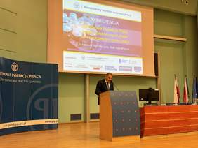 Pokaż zdjęcie: Mężczyzna przemawia z podium na konferencji w sali wykładowej. W tle widoczny jest ekran z prezentacją zatytułowaną „Państwowa Inspekcja Pracy wobec dynamicznych zmian na współczesnym rynku pracy”, flagi oraz baner Państwowej Inspekcji Pracy.