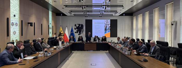 Pokaż zdjęcie: Spotkanie w nowoczesnej sali konferencyjnej. Przy dużym stole siedzą osoby w formalnych strojach, a na ekranie wyświetlana jest prezentacja. W tle widoczne są flagi Polski, Unii Europejskiej i regionu z herbem czarnego orła.