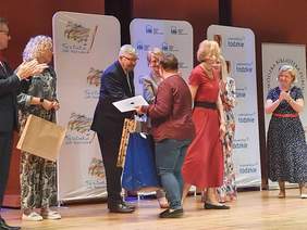 Pokaż zdjęcie: Na scenie stoją osoby. Pan podaje rękę pani i uśmiecha się do kobiety. Stojący obok ludzie biją brawo. 