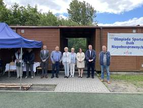 Pokaż zdjęcie: Przed drewnianym budynkiem na kostce brukowej stoi 9 osób. Mężczyźni ubrani w marynarki Kobiety ubrane również marynarki i odzież sportową. Wśród tych osób znajduje się ksiądz.