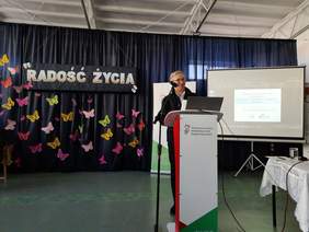 Pokaż zdjęcie: Przy mównicy stoi uśmiechnięty prelegent w okularach.