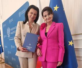 Pokaż zdjęcie: Dwie uśmiechnięte kobiety pozują do zdjęcia. Jedna z nich trzyma w rękach teczkę z logo OHP. Za nimi flagi Unii Europejskiej i Polski. W tle ścianka promocyjna Województwa Małopolskiego