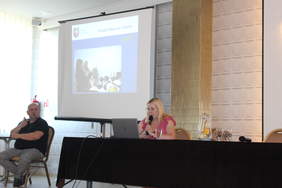 Pokaż zdjęcie: W sali konferencyjnej za stołem prezydialnym siedzi z mikrofonem, z lewej strony duży ekran z wyświetlaną prezentacją, po lewej stronie przed prezentacją siedzi tłumacz języka migowego