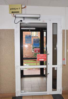 Pokaż zdjęcie: Szklane drzwi wewnętrzne z naklejką: „drzwi automatyczne”. Nad drzwiami wewnętrznymi przytwierdzona jest tabliczka z napisem: „pokój nr 7”. Z kolei na drzwiach zewnętrznych widnieje napis: „pchać”, jako również przymocowane zostały dwa plakaty.