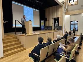 Pokaż zdjęcie: Na scenie za mównicą stoi mężczyzna i mówi do mikrofonu. Za nim wielki telebim z prezentacją. Na dole rzędy jasnych foteli, na których siedzą ludzie.