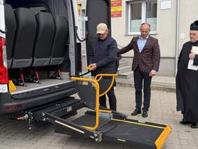 Pokaż zdjęcie: Mężczyzna obsługuje windę dla osób z niepełnosprawnościami w busie. Obok stoją dwaj mężczyźni przed budynkiem Urzędu Gminy Linia.