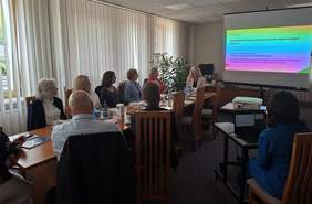 Pokaż zdjęcie: W sali grupa osób siedzi przy długim stole, głównie kobiety w eleganckich strojach. Mężczyzna siedzi tyłem. Na końcu stołu znajduje się ekran, na którym wyświetlana jest prezentacja.