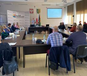 Pokaż zdjęcie: Sala konferencyjna, stoły ustawione w podkowę, po lewej stronie stoi baner z hasłem powiat włodawski, przy stole siedzą słuchacze, mężczyzna prelegent prowadzi szkolenie