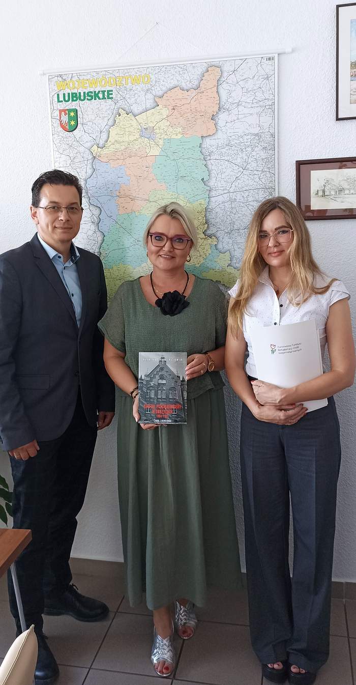 Pokaż zdjęcie: Obok siebie na tle mapy województwa lubuskiego stoją dwie kobiety spośród których jedna trzyma książkę w ręku, a druga teczkę z logo PFRON, a obok w garniturze stoi mężczyzna