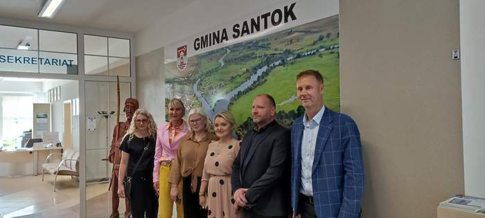 Pokaż zdjęcie: Uczestnicy spotkania ubrani odświętnie stoją na tle ścianki promocyjnej w Urzędzie Gminy Santok