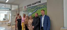 Pokaż zdjęcie: Uczestnicy spotkania ubrani odświętnie stoją na tle ścianki promocyjnej w Urzędzie Gminy Santok
