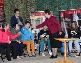 Pokaż zdjęcie: Na scenie stoją krzesełka na których siedzą dzieci. Przed nimi stoi kobieta i trzyma w ręku mikrofon.  Wysuwa rękę z mikrofonem w kierunku jednego z dzieci.