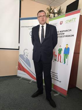 Pokaż zdjęcie: Mężczyzna w garniturze pozuje na tle baneru z logo OWiT.