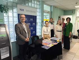 Stanowisko informacyjne NFZ i PFRON w budynku publicznym; kilka osób stoi przy stoliku z ulotkami i materiałami edukacyjnymi, w tle bannery instytucji oraz automat biletowy ZUS.