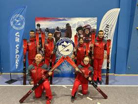 6 narciarzy, kobiety i mężczyźni w klubowych czerwonych strojach pozują do zdjęcia. Za nimi niebieskie tło.