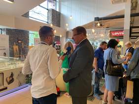 Pokaż zdjęcie: Dwóch mężczyzn stoi na tle wystawy obrazów w galerii. Jeden z nich trzyma mikrofon, drugi udziela wywiadu.