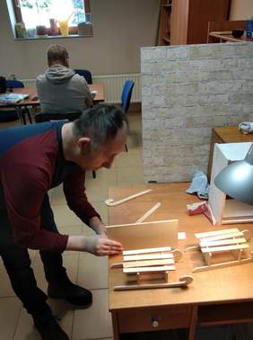 Pokaż zdjęcie: W pomieszczeniu jedna osoba siedzi tyłem przy biurku pod oknem druga osoba mężczyzna stoi pochylony nad biurkiem i trzyma deseczkę obok stoi dwoje miniaturowych sanek