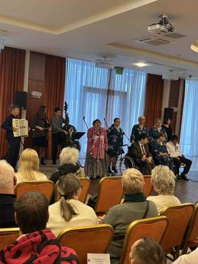 Pokaż zdjęcie: W sali odbywa się koncert. Niektórzy śpiewają, a inni grają na instrumentach muzycznych. Osoby na widowni oglądają występ. 