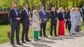 Pokaż zdjęcie: Uczestnicy wydarzenia stoją w szeregu podczas oficjalnej części obchodów Dnia Godności. Jedna z kobiet trzyma mikrofon i przemawia. Obok niej znajdują się lokalni samorządowcy, członkowie organizacji i goście. W tle widoczne są plansze prezentujące działalność placówek wspierających osoby z niepełnosprawnością.