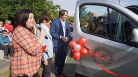 Pokaż zdjęcie: Uroczystość przed Szkołą Podstawową Specjalną nr 19 w Kaliszu. Od lewej stoją trzy osoby: kobieta w pomarańczowej koszuli przemawiająca do mikrofonu, kobieta w jasnej marynarce oraz mężczyzna w granatowym garniturze. Po prawej stronie widoczny jest srebrny bus przystrojony czerwonymi balonami i wstęgą. Na drzwiach pojazdu znajduje się okrągłe logo szkoły z rysunkiem dłoni nałożonych na siebie. W tle grupa zgromadzonych ludzi oraz drzewa.
