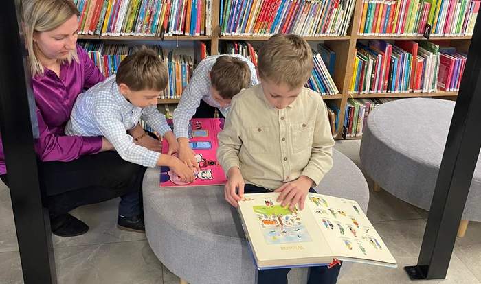 Pokaż zdjęcie: Troje dzieci siedzi przy stole i wspólnie ogląda książkę, obok stoi dorosła osoba wspierająca ich w czytaniu; w tle regały wypełnione kolorowymi książkami w bibliotece.