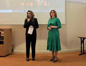 Pokaż zdjęcie: Na wzorzystym dywanie stoją dwie kobiety w okularach. Trzymają mikrofony. Jedna jest ubrana w spodnie i marynarkę mówi do mikrofonu. Druga jest ubrana w jednolitą sukienkę. Za nimi wisi telebim z odtwarzaną prezentacją.